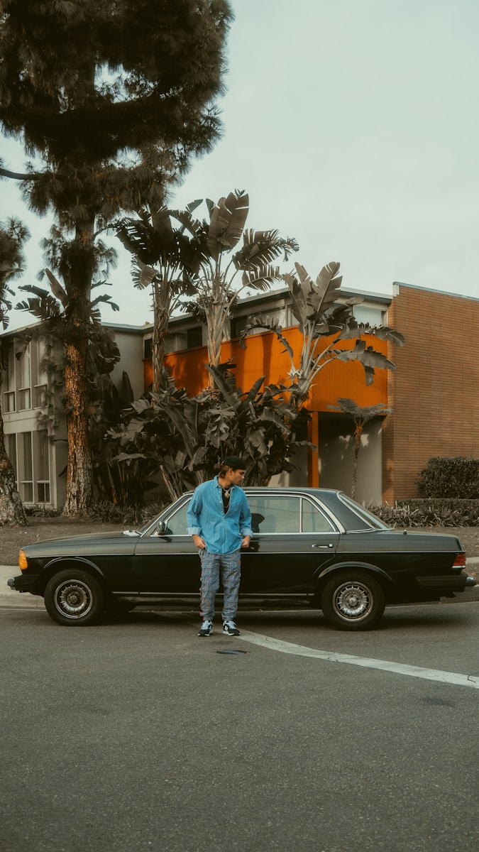 man in blue jacket and blue denim jeans standing beside black coupe with auto insurance during daytime